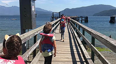 Children wearing backpacks walk along a wooden pier beside the ocean during a summer camp outing.