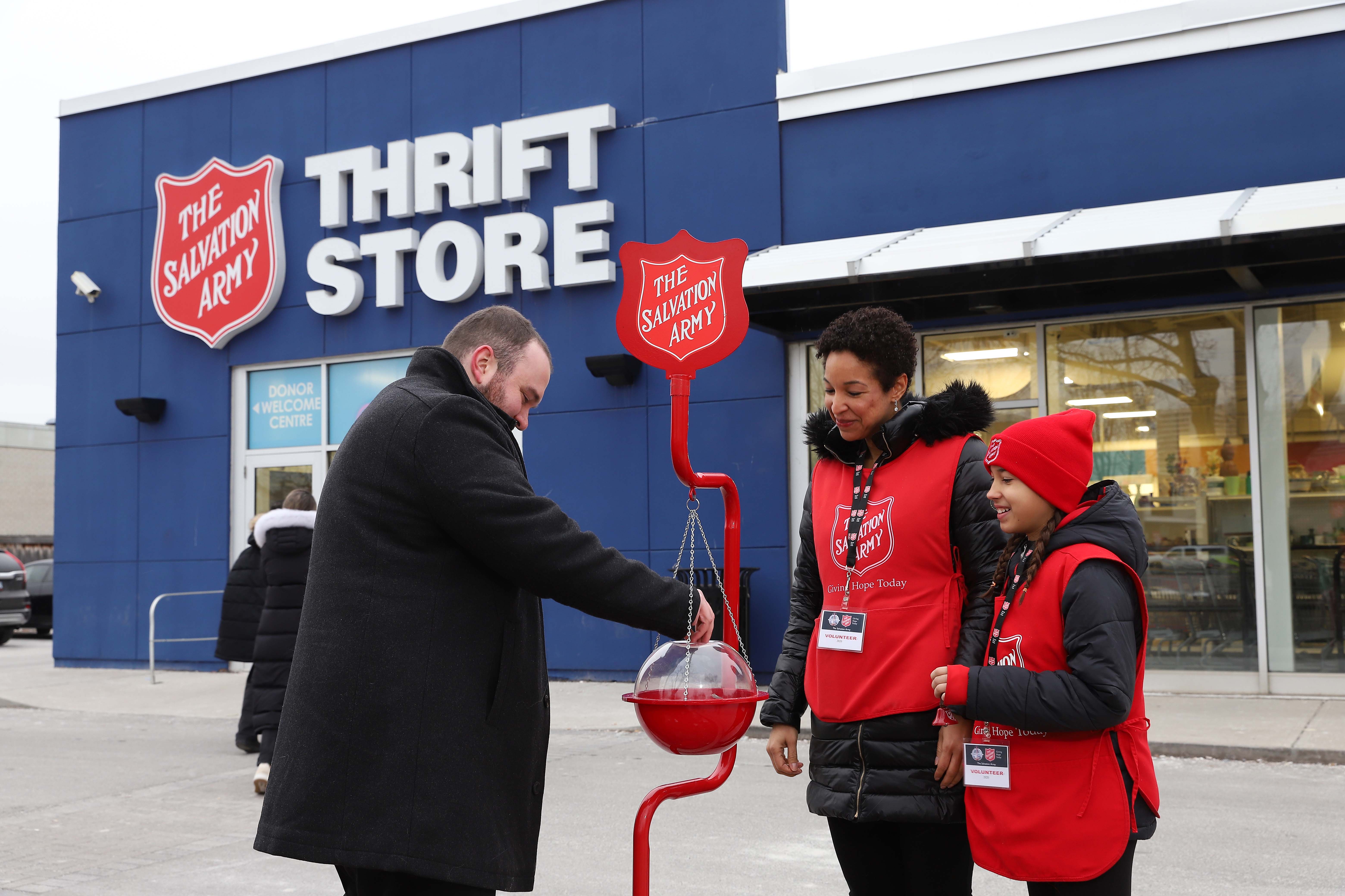 Two Salvation Army volunteers stand beside a red Christmas kettle outside a thrift store as a man places a donation into the kettle during the Christmas Kettle Campaign.