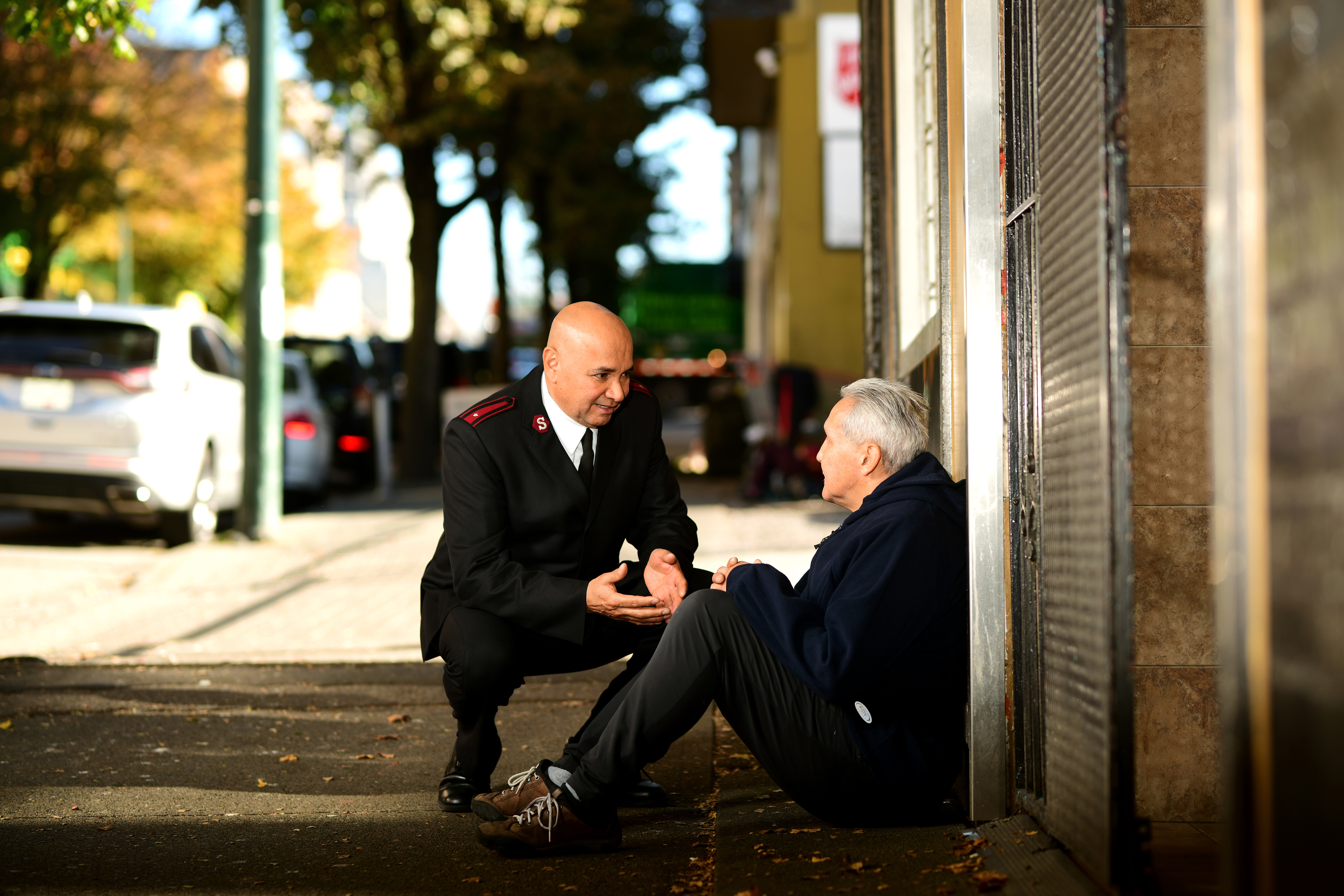 A Salvation Army officer kneels on a sidewalk speaking with a seated older man outside a building.
